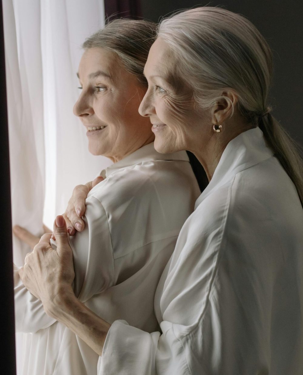 Two elderly women in white robes embracing near a window, symbolizing love and companionship.